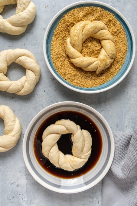 One ring shaped simit dough soaked in molasses in a bowl, another in a bowl of toasted sesame seeds and more braided simits on the side for this process.