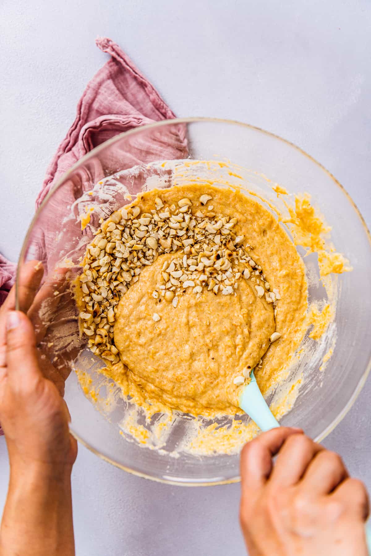 Hands stirring pumpkin muffin batter and chopped peanuts with a spatula in a mixing bowl.