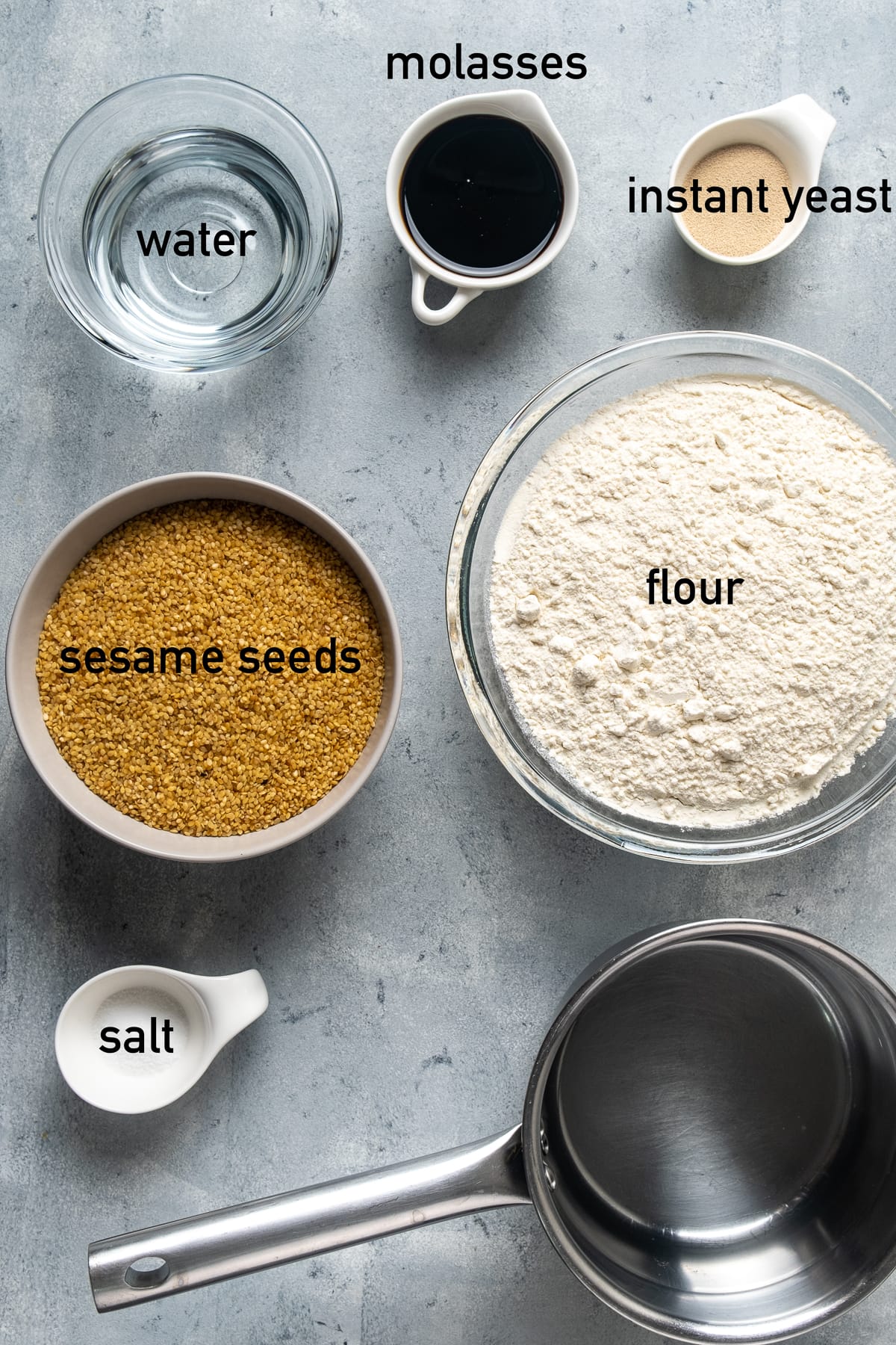 Sesame seeds, flour, grape molasses, instant yeast, water and salt all in separate bowls on a grey background.