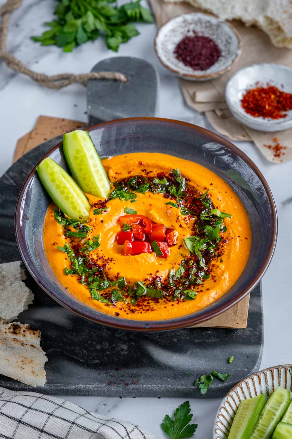 Red pepper hummus served with cucumber sticks, garnished with parsley and spices.