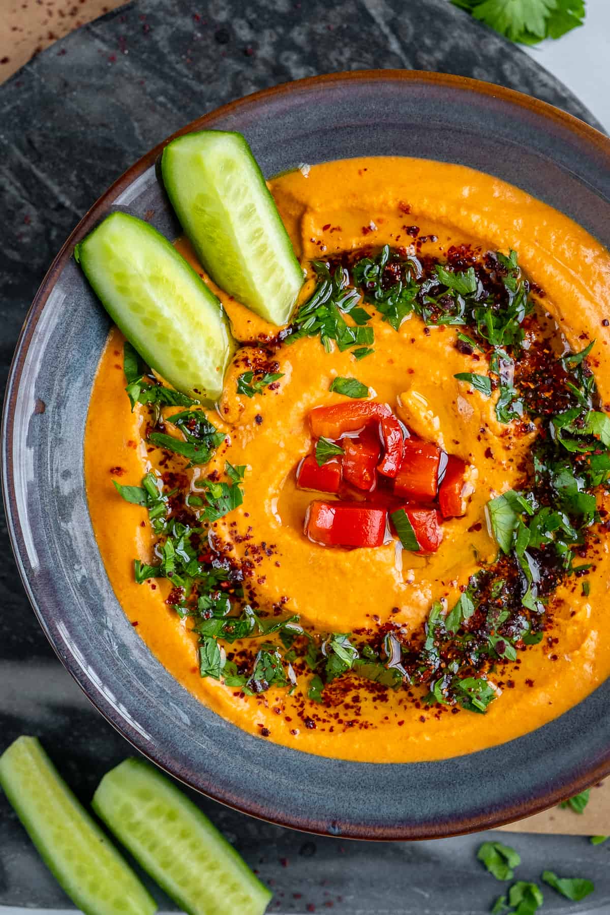 Red pepper hummus garnished with parsley, red pepper flakes, sumac, chopped roasted peppers and cucumber sticks.