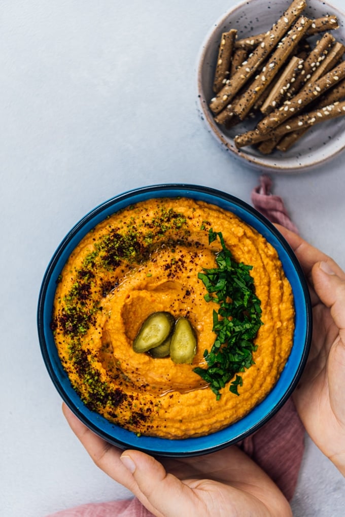 Woman serving roasted red pepper hummus in a blue bowl garnished with parsley, ground pistachio, sumac and dill pickles accompanied by crackers.