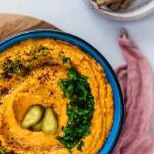 Roasted red pepper hummus in a blue bowl garnished with parsley, ground pistachio, sumac and dill pickles accompanied by crackers.