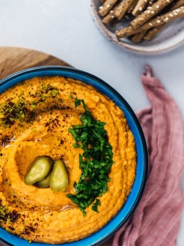 Roasted red pepper hummus in a blue bowl garnished with parsley, ground pistachio, sumac and dill pickles accompanied by crackers.