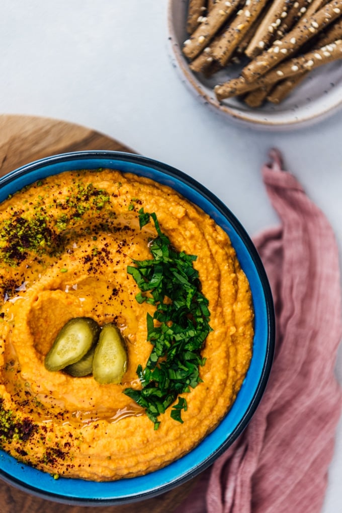 Roasted red pepper hummus garnished with parsley, sumac, ground pistachio and pickles in a blue bowl