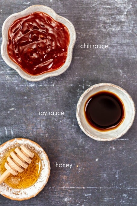 Chili sauce, soy sauce and honey in separate bowls on a dark background.