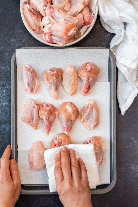 Hands pat drying chicken wings on a baking sheet using paper towel.