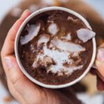 Holding a cup of almond milk hot chocolate topped with coconut oil and coconut flakes.