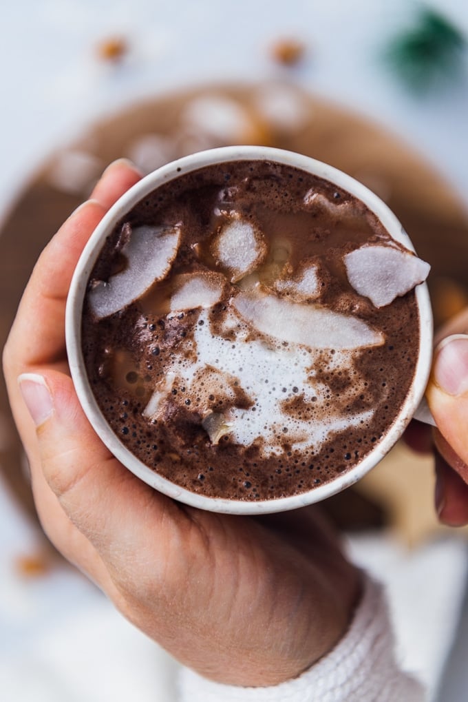 Holding a cup of almond milk hot chocolate topped with coconut oil and coconut flakes.