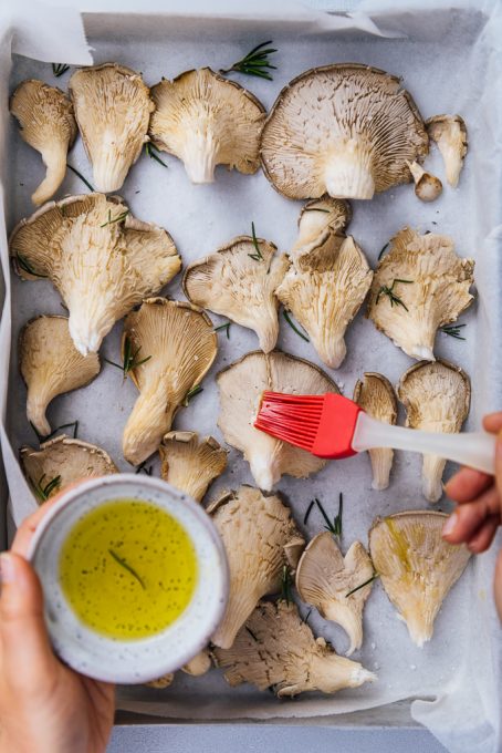 Hands brushing oyster mushrooms with olive oil in a baking sheet.
