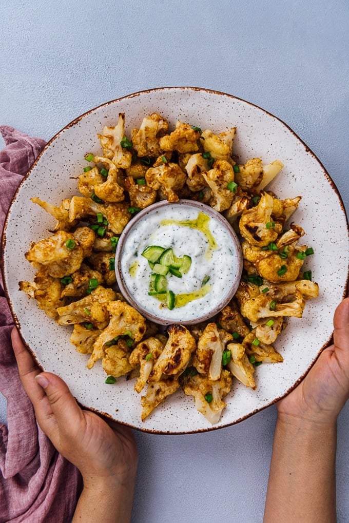 Hands holding roasted spicy cauliflower wings in a ceramic bowl served with a small bowl of yogurt cucumber dip.