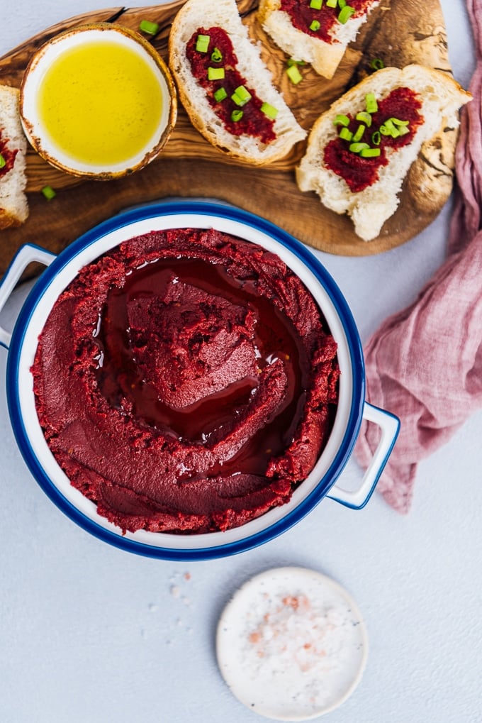 Tomato paste in a white container accompanied by slices of bread topped with tomato paste and herbs on a wooden board, a little salt on a tiny ceramic plate.