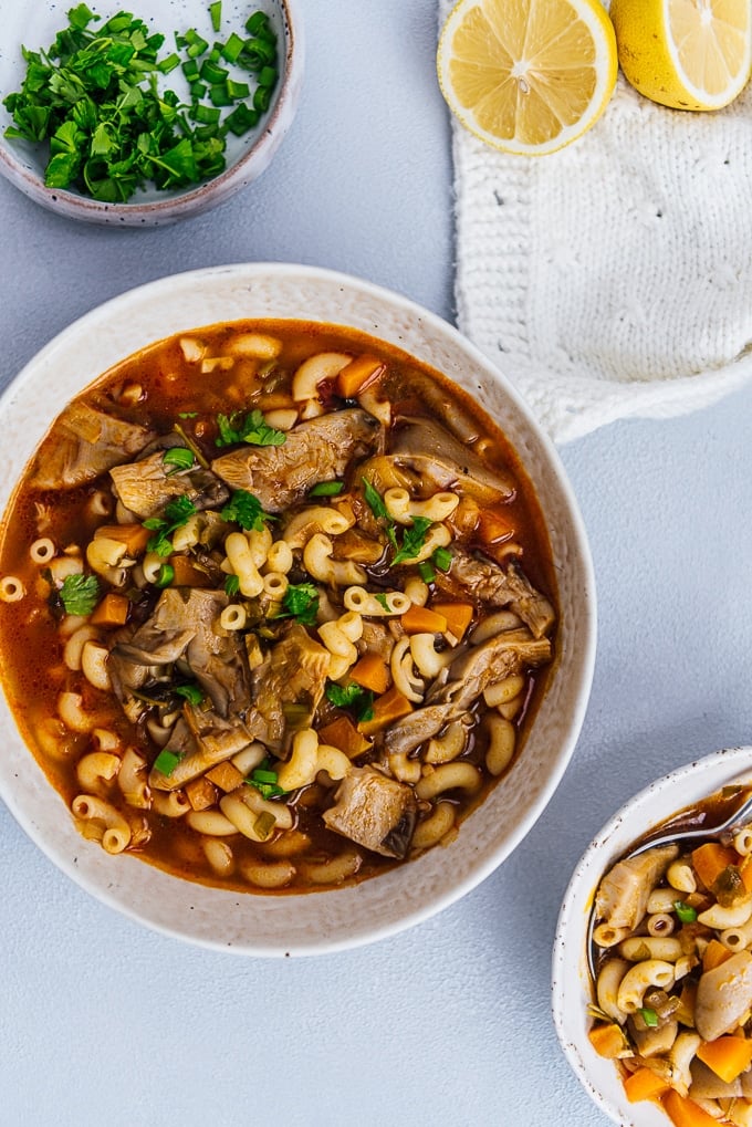 Oyster mushroom soup with pasta garnished with herbs in a bowl, lemon and herbs accompany.