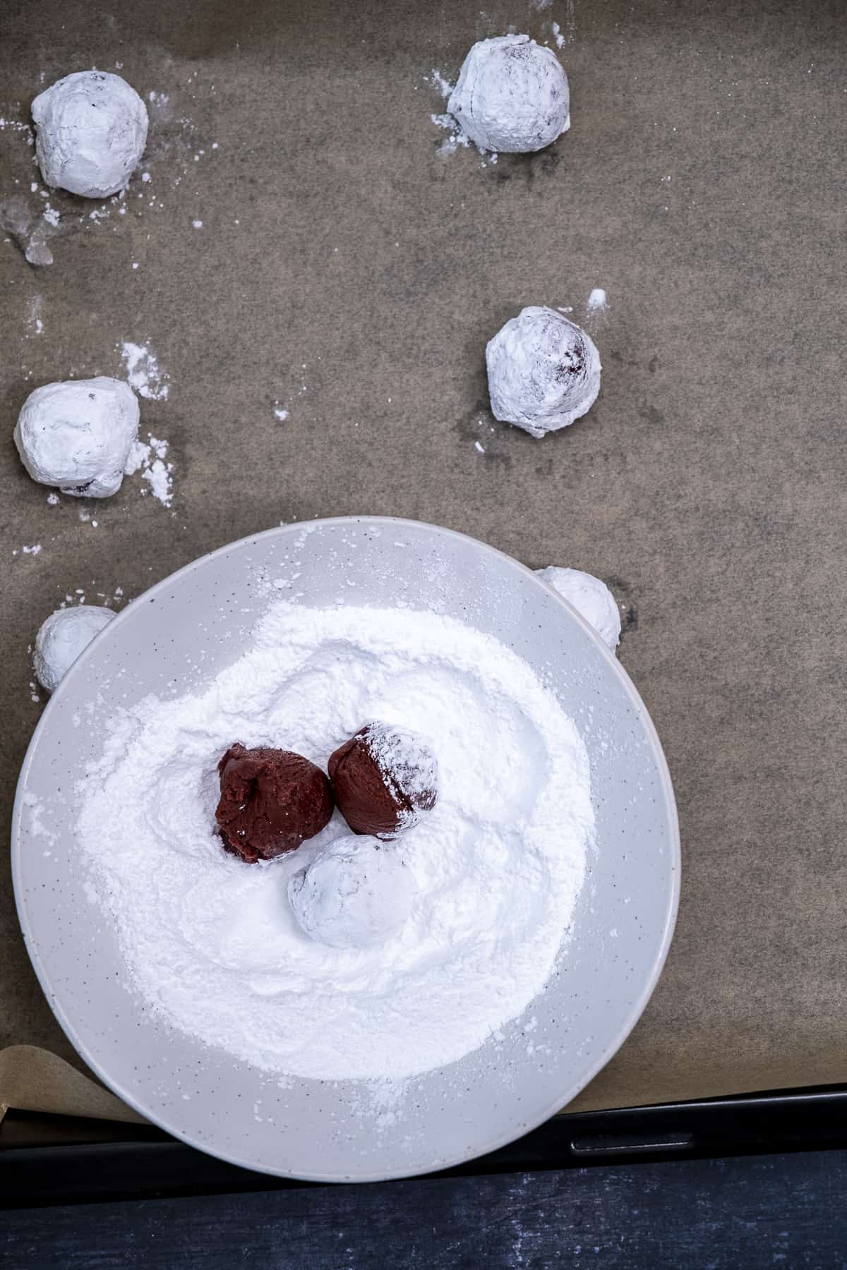 Red velvet cookie dough balls in a bowl of powdered sugar and more cookie dough balls around it on a baking sheet.