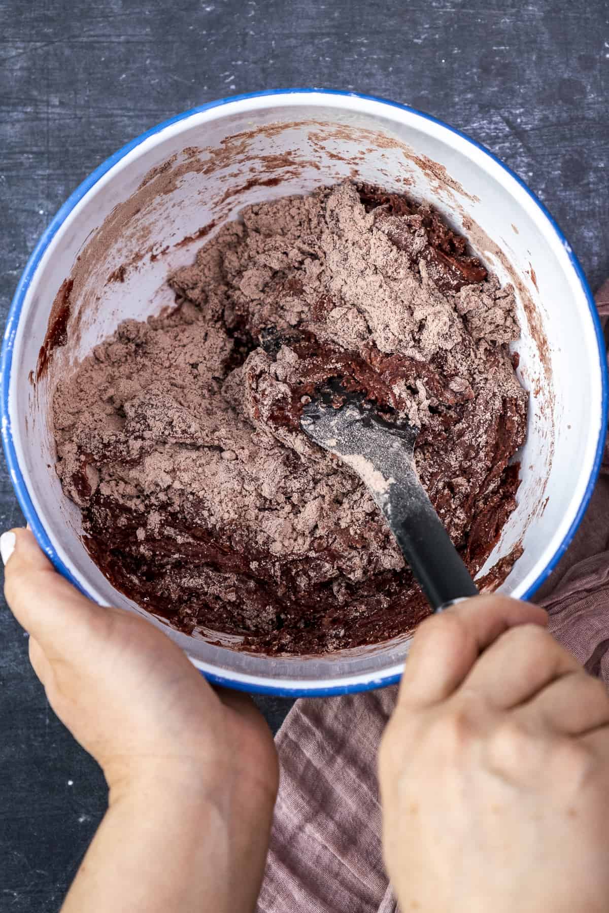 Hands mixing red velvet cookie dough with a black spatula in a white bowl.