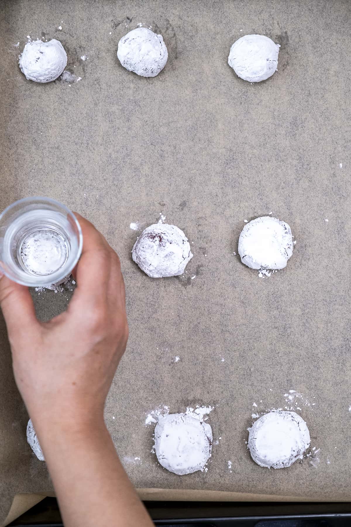 A hand flattening the cookie dough balls on a baking sheet using a glass cup.