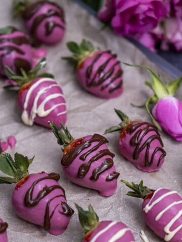 Pink chocolate coated strawberries with dark and white chocolate drizzles on baking paper and pink roses behind them.