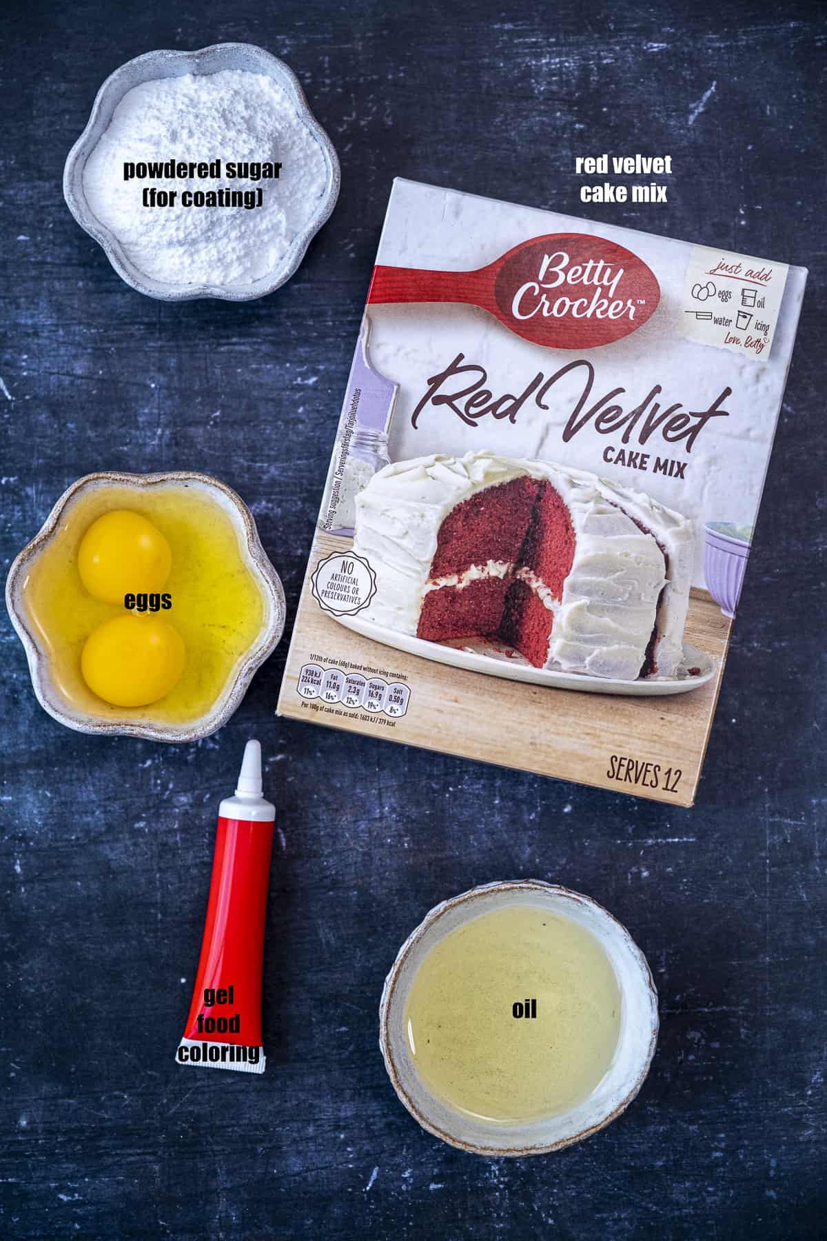A box of red velvet cake mix, eggs, powdered sugar, oil and a tube of red food coloring on a dark background.