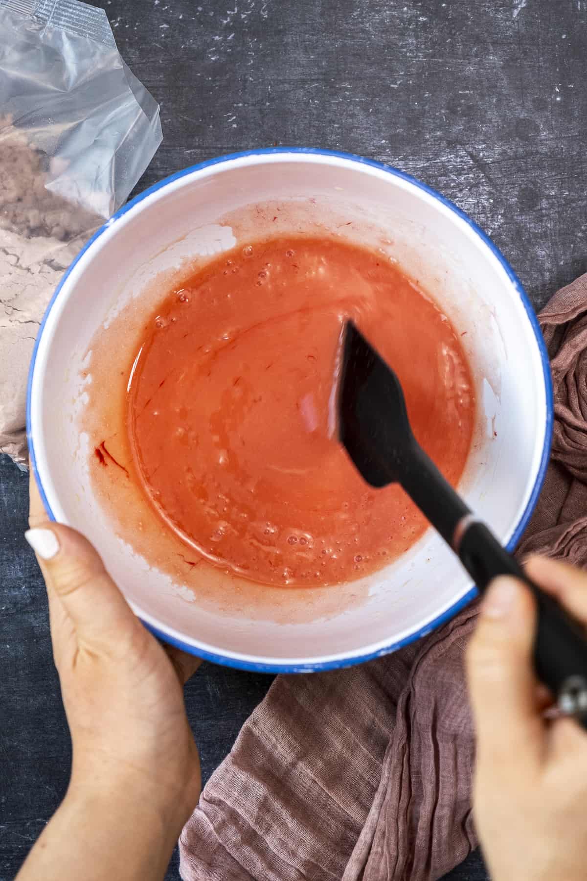 Hands mixing eggs, oil and red food coloring with a spatula in a white bowl.