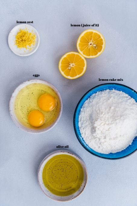 Lemon zest, eggs, halved lemon, lemon cake mix, olive oil in separate bowls on a light background.