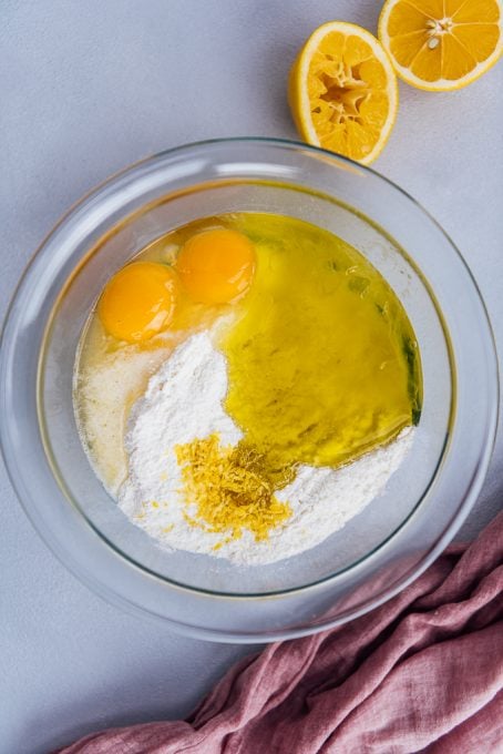 Eggs, flour, lemon zest, oil in a glass mixing bowl, lemons on the side.