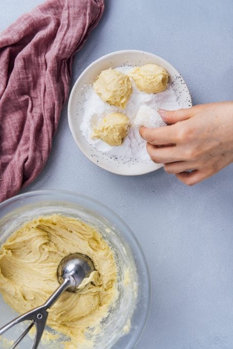 A hand rolling lemon cookie balls into powdered sugar in a bowl, cookie dough on the side.