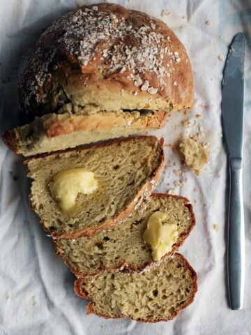 A round no yeast bread on a white kitchen towel, half sliced, some slices topped with butter and a knife on the side.