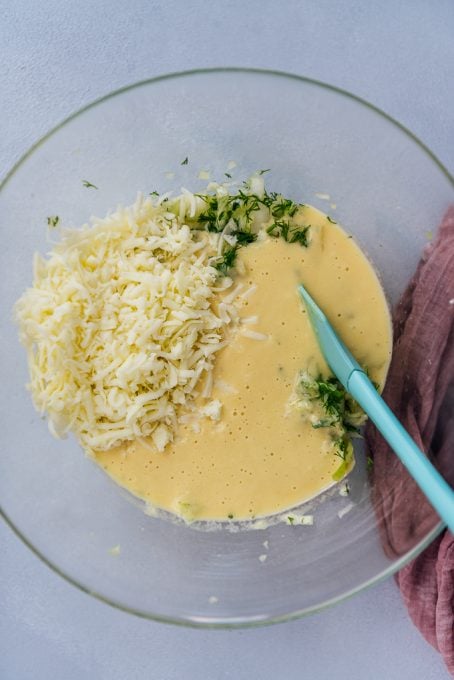 A mixture of egg batter, shredded cheese, shredded cabbage and fresh dill in a glass bowl.