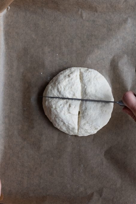 A hand making a cross on a round bread dough with a knife.