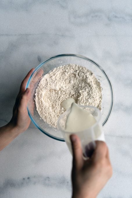 Hands pouring yogurt into a flour mixture in a large glass bowl.