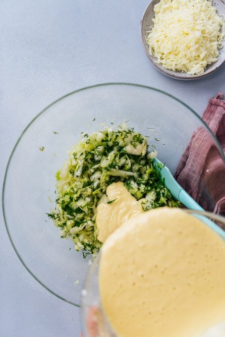 Hands pouring the wet batter over shredded cabbage and fresh dill in a glass mixing bowl.