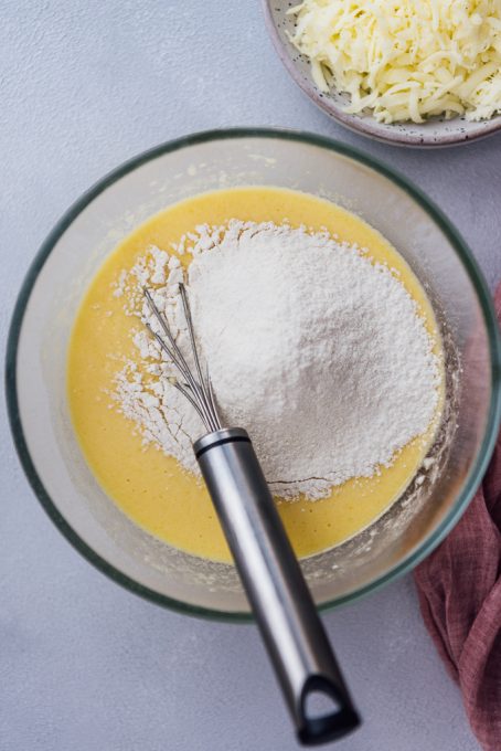 Preparing a batter with eggs, yogurt, oil and flour in a glass bowl, shredded cheese on the side.