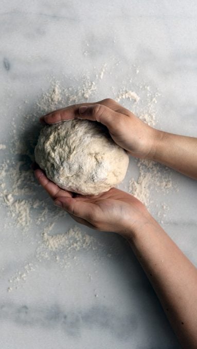 Hands rolling a bread dough into a ball on a marble counter.