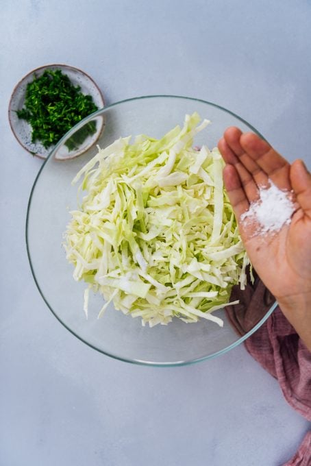 Hand sprinkling salt over shredded cabbage in a glass bowl.