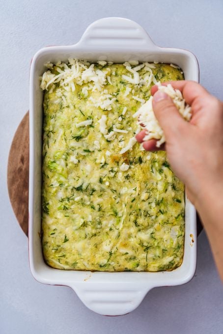 A hand sprinkling shredded cheese over the baked cabbage casserole in a rectangular white baking pan.