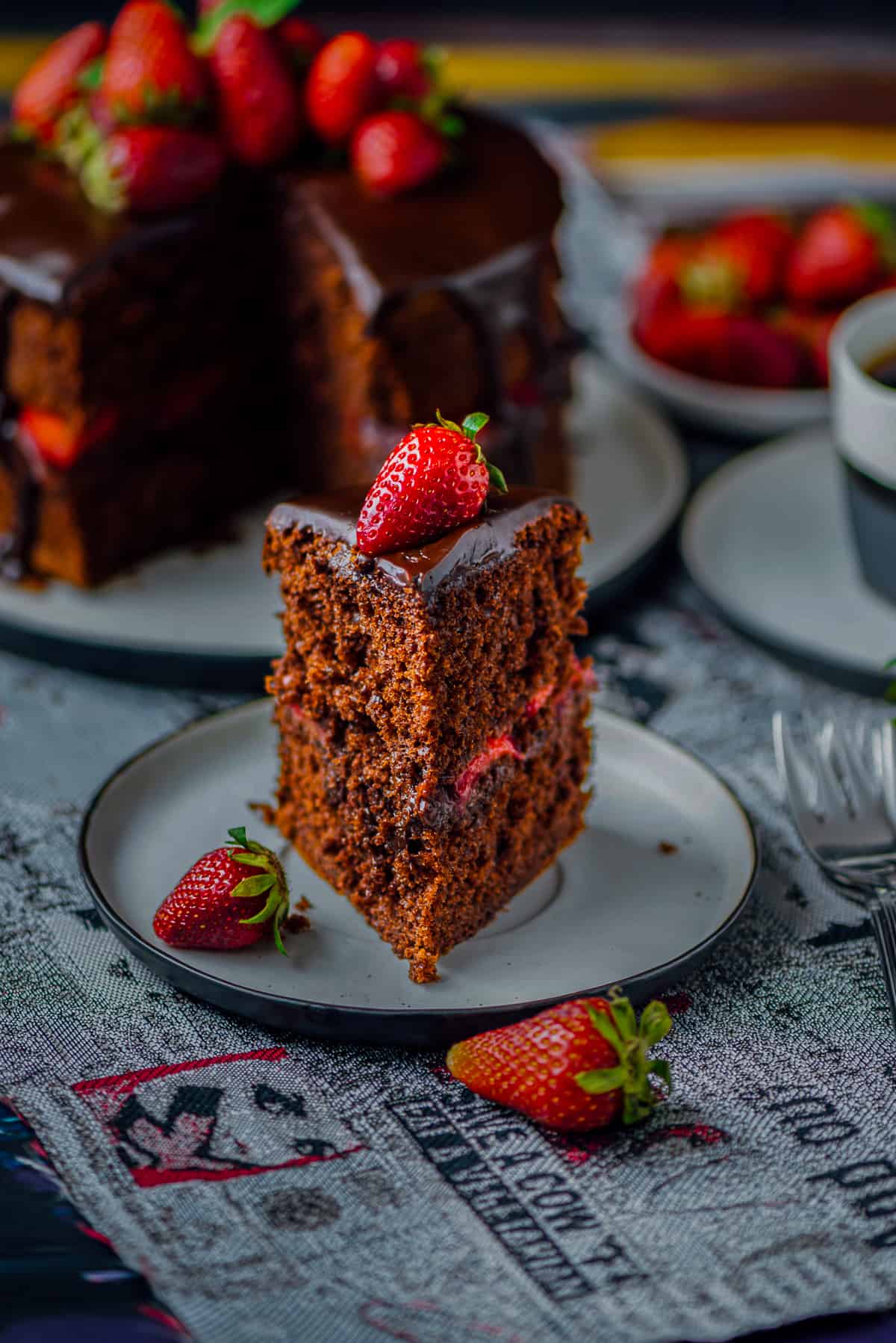 A slice of chocolate strawberry cake on a plate, the whole cake, strawberries and a coffee cup behind it.