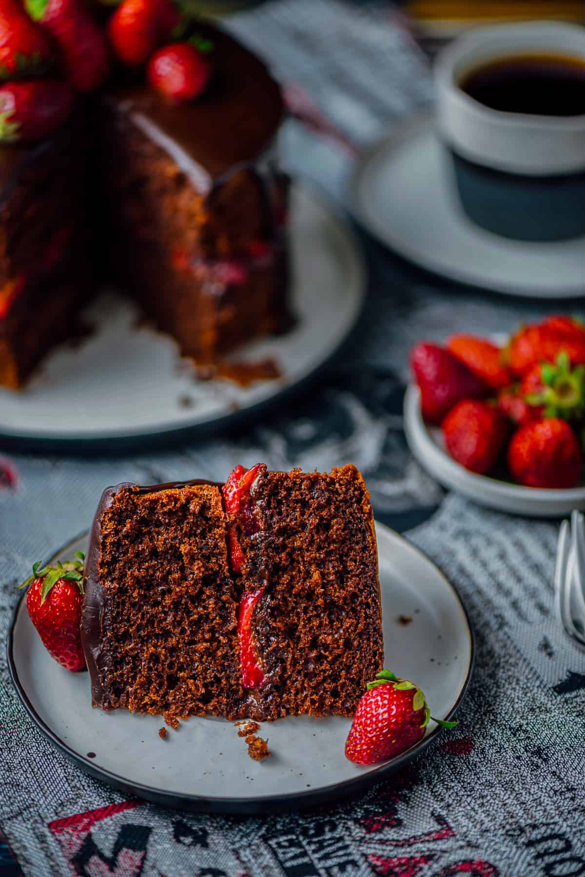 A slice of chocolate cake with strawberries served on a plate and the whole cake, more strawberries and a cup of coffee behind it.