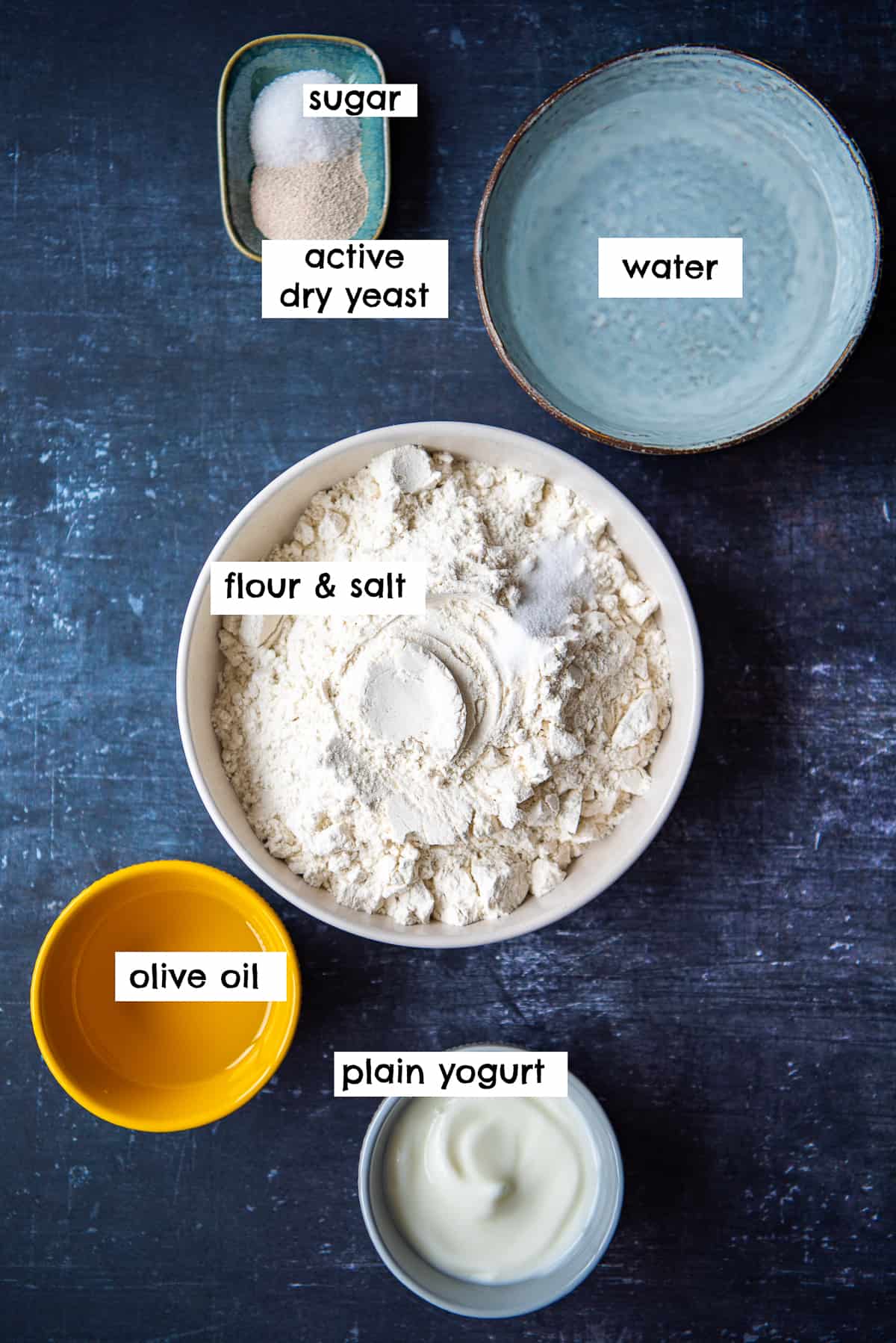 Sugar, yeast, water, flour, Greek yogurt and olive oil photographed on a dark background.