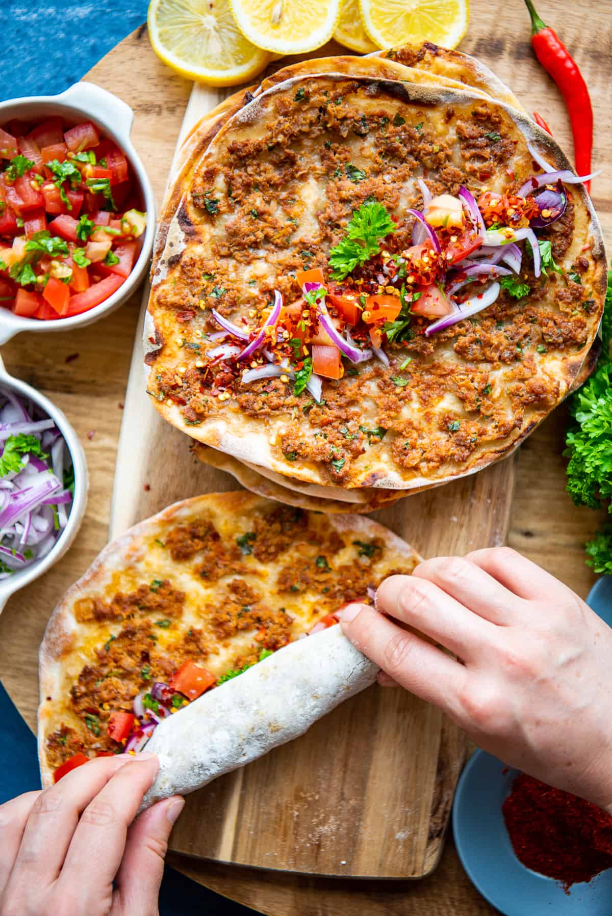 Hands making lahmacun wrap, sliced onions and tomato salad on the side.