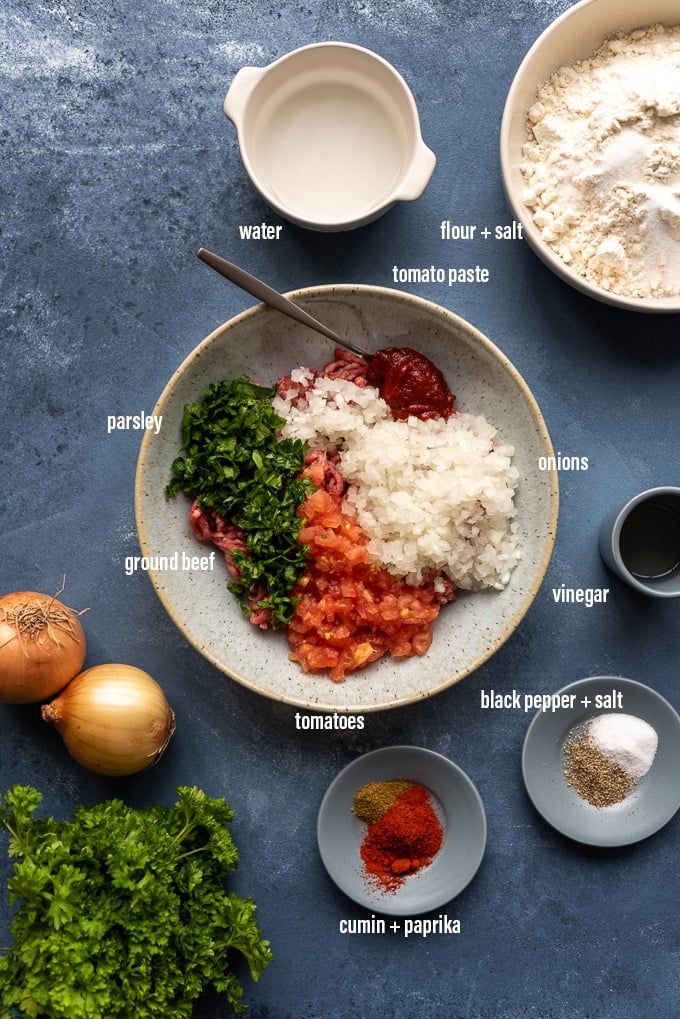 Lahmajun recipe ingredients including ground beef, onions, tomatoes, tomato paste, parsley, spices, vinegar, flour and salt on a dark grey background.