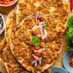 Lahmacun served on a wooden board, one topped onions, tomatoes and parsley.