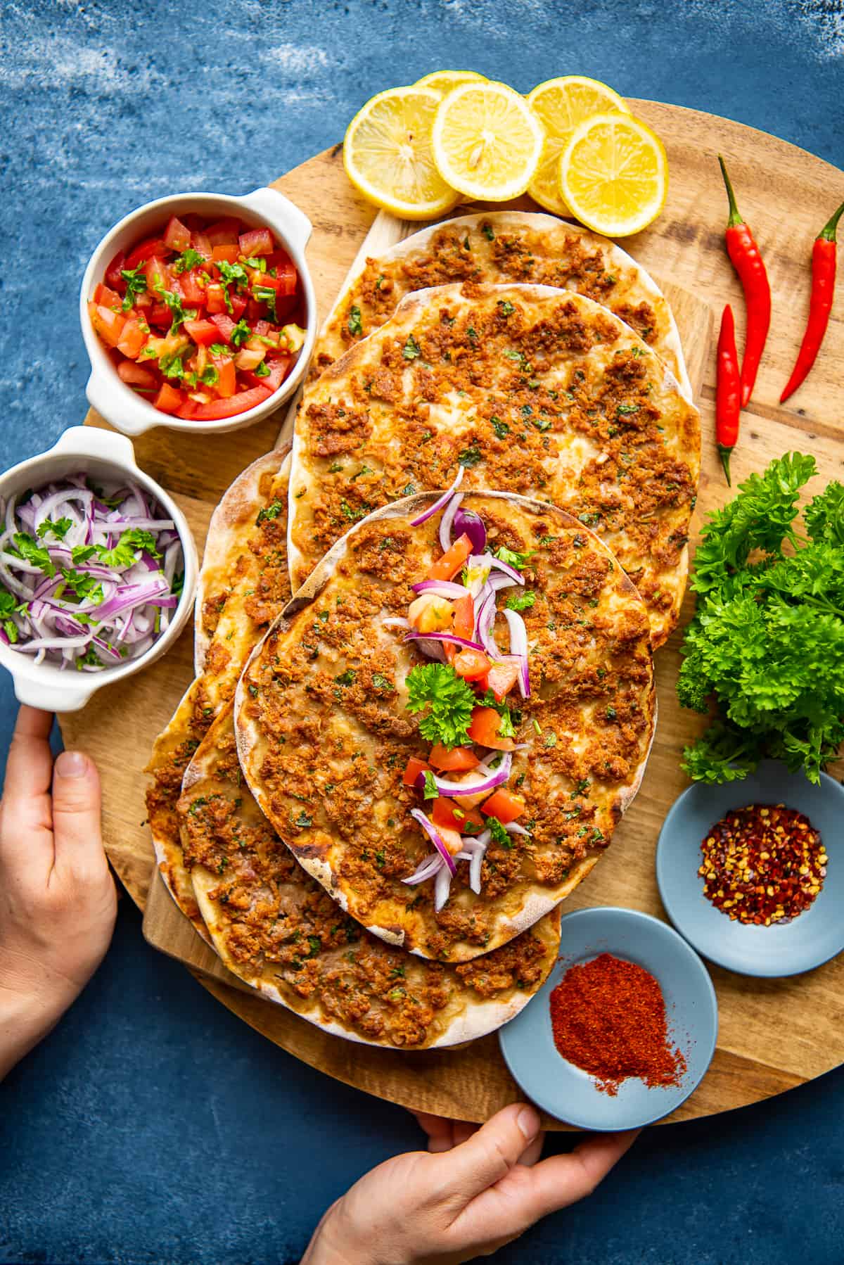 Hands holding a wooden board with lahmacun, salads and spices on it.
