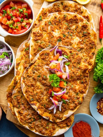 Hands serving lahmacun, sliced onion, tomato salad and spices on a wooden board.