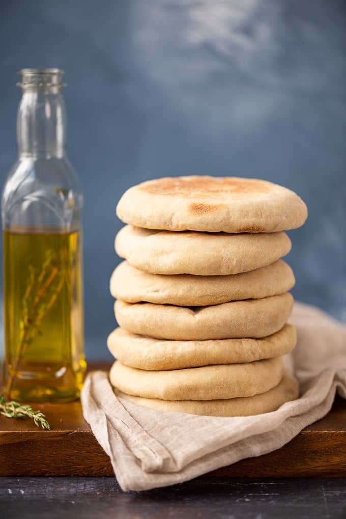 A stack of Turkish flatbread called bazlama on a wooden board, a bottle of olive oil and fresh thyme accompany.