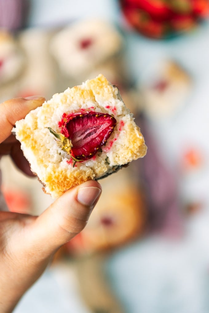 A hand holding a slice of coconut ice with a strawberry topping photographed closely from top view.