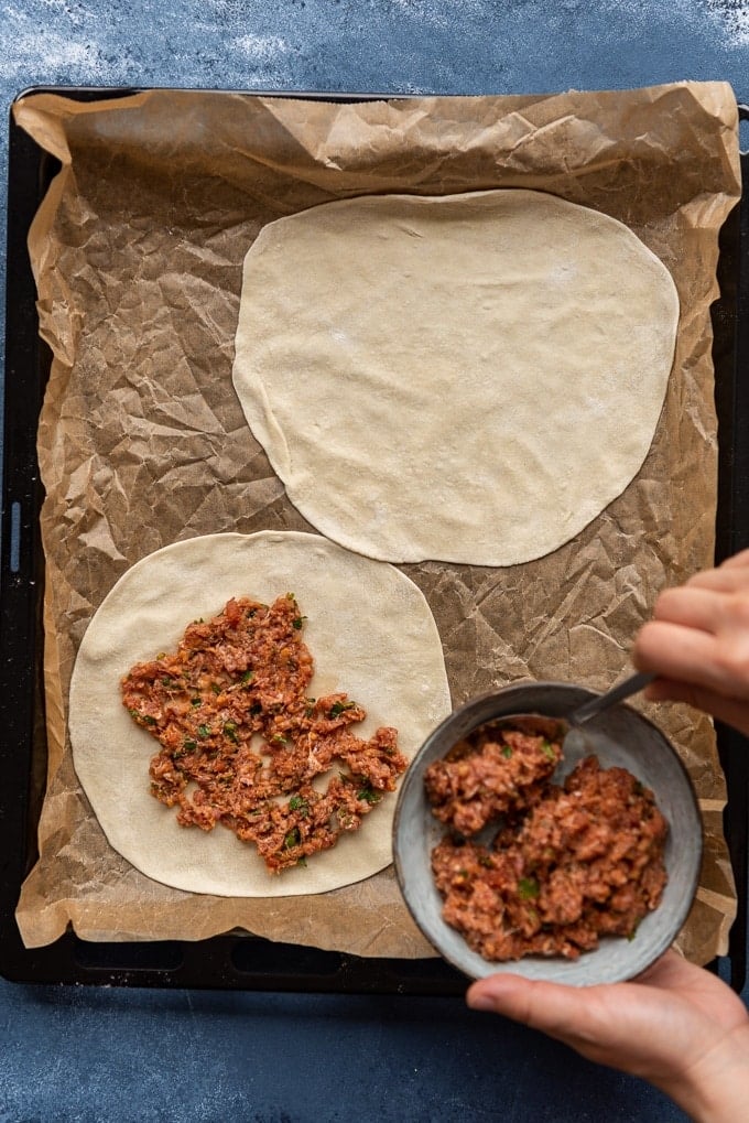 Woman making lahmacun from scratch with homemade dough and ground beef filling on a parchment paper lined baking sheet.