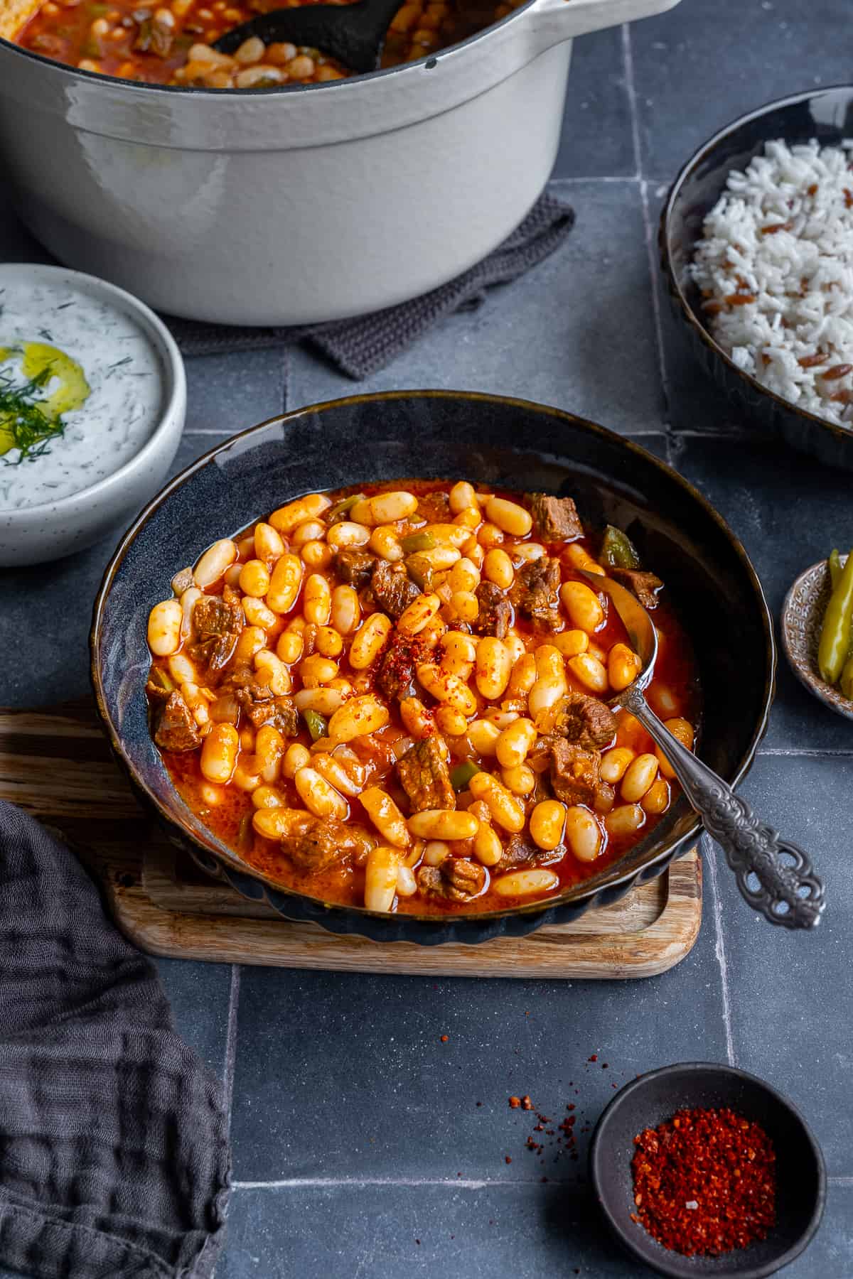 Turkish bean stew in a dark colored bowl with a spoon in it, a pot of more stew, cacik, rice pilav, pickles and red pepper flakes accompany.