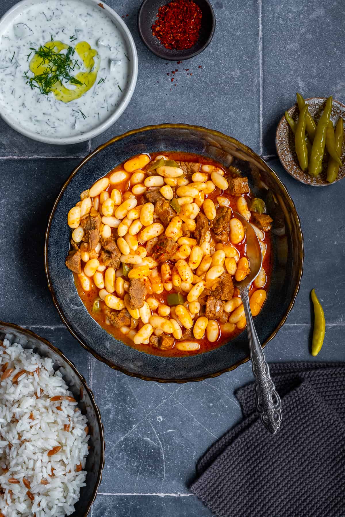 Turkish bean stew in a dark colored bowl with a spoon in it from top view, rice pilav, cacik and pickles accompany.