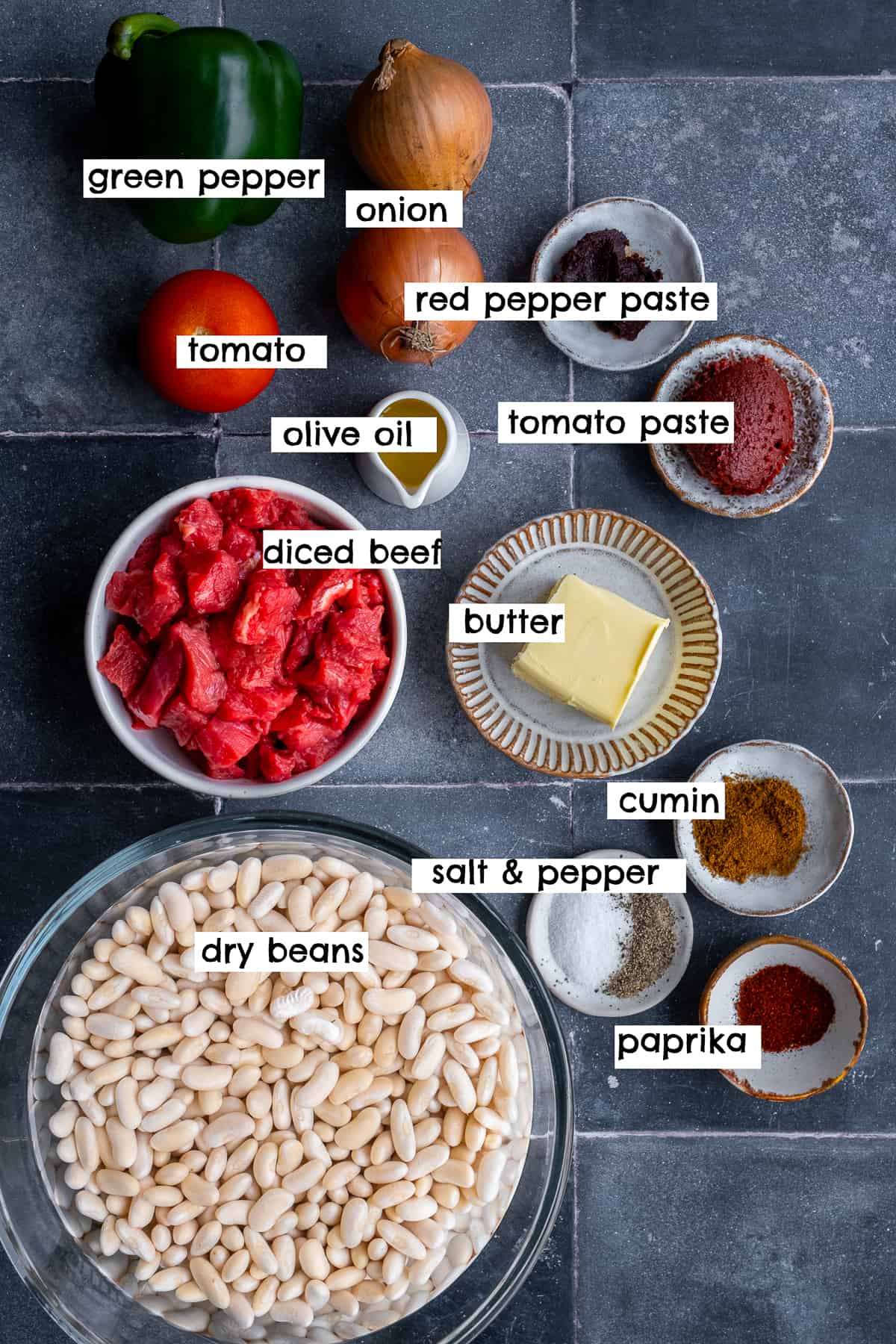 Soaked white beans, diced beef, butter, olive oil, tomato paste, pepper paste, onions, tomato, green bell pepper, paprika, cumin, salt and pepper photographed from top view.