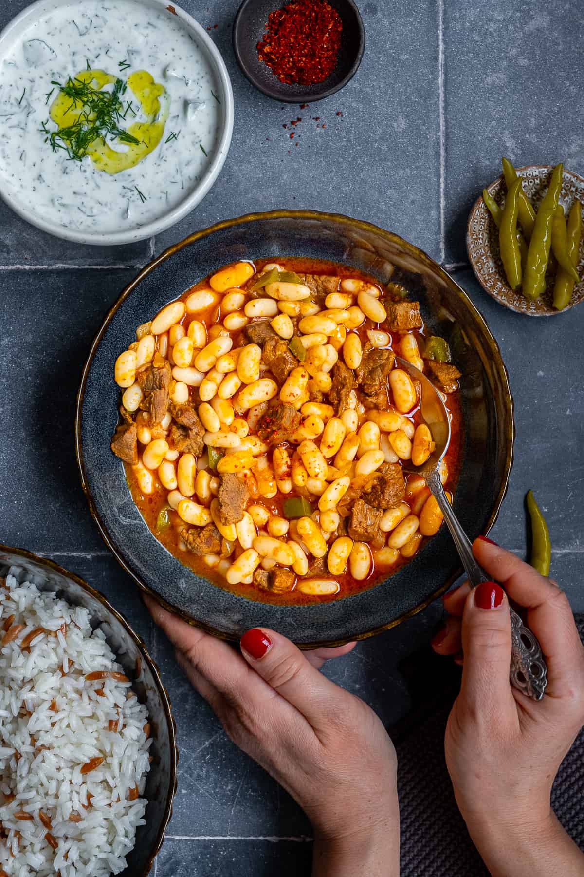 Woman about to eat Turkish beans in a dark colored bowl with a spoon and cacik, rice pilav and pickles accompany.